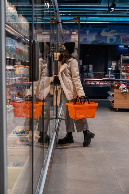 Mujer en abrigo y mascarilla médica en la tienda de abarrotes abriendo la puerta del frigorífico