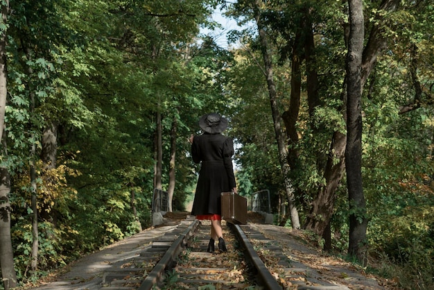 Mujer con abrigo negro y sombrero de estilo años 40 con valija retro en las manos camina a lo largo de rieles atravesando el bosque Vista posterior