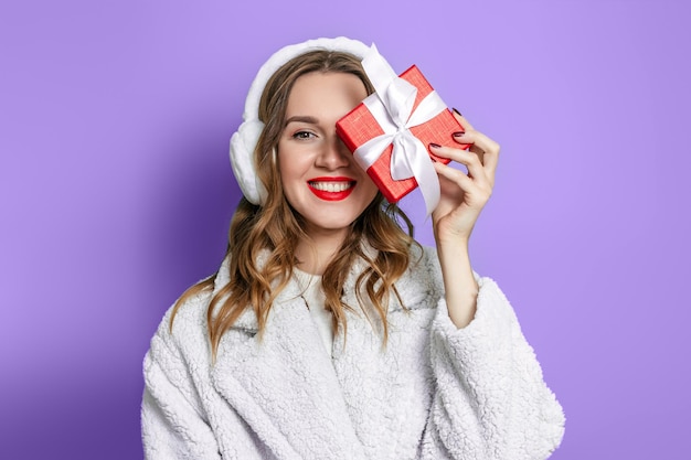 Mujer en un abrigo de piel blanco y auriculares de piel con una caja de regalo sonríe aislado sobre un fondo púrpura