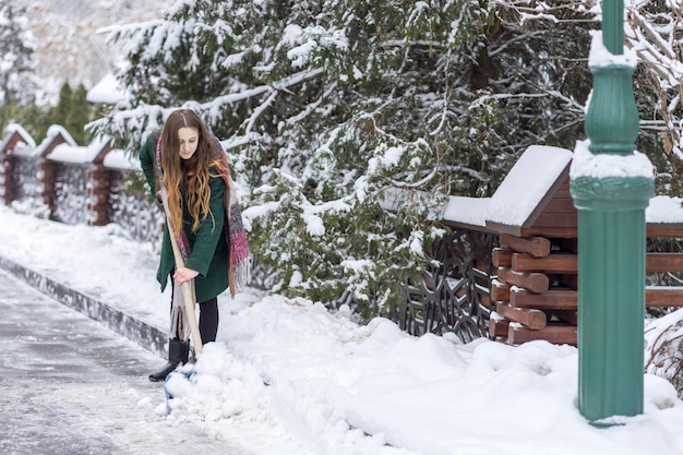 mujer con un abrigo verde limpia la carretera en un parque de la ciudad de la nieve