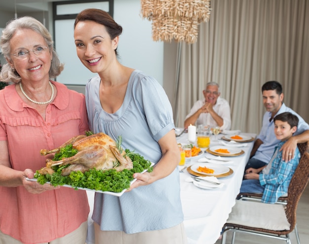 Mujer y abuela sosteniendo pollo asado con la familia en la mesa de comedor