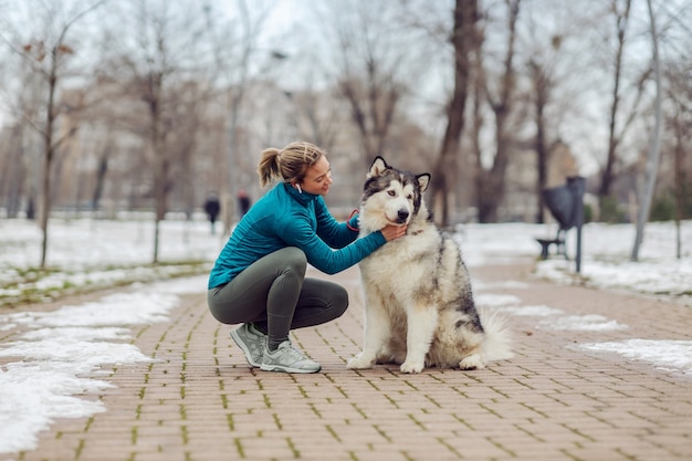 Mujer acariciando a su perro mientras se agacha en un parque público en un día nevado de invierno. Mascotas, actividades de fin de semana, invierno, nieve, amistad