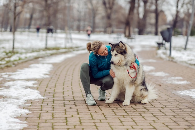 Mujer acariciando a su perro mientras se agacha en un parque público en un día nevado de invierno. Mascotas, actividades de fin de semana, invierno, nieve, amistad