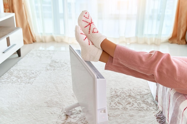 Una mujer acogedora que usa calcetines de punto suaves y cómodos se calienta los pies con un moderno calentador eléctrico portátil blanco en casa