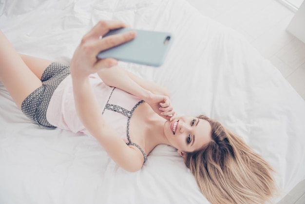 Mujer acostada en la cama en casa con smartphone tomando selfie