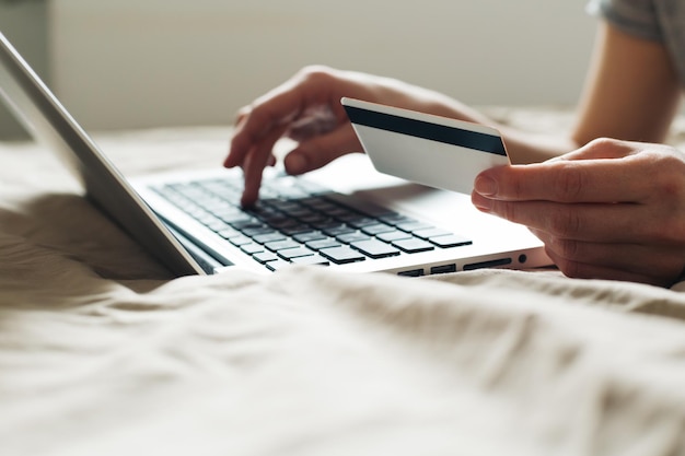 Mujer acostada en una cama comprando en línea en una computadora portátil usando un primer plano de tarjeta de crédito