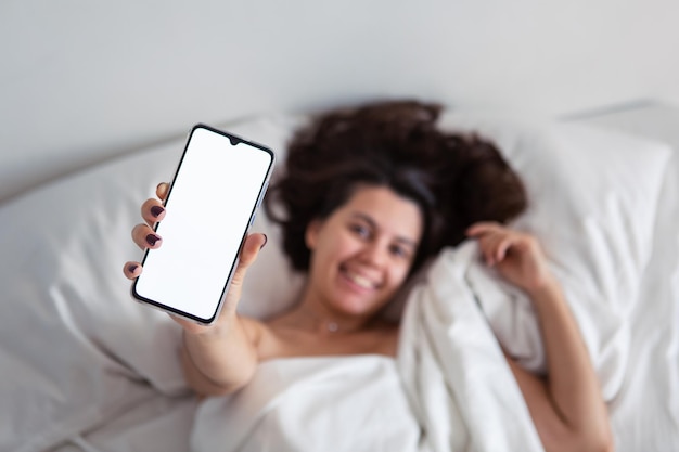 Mujer acostada en la cama mostrando teléfono con pantalla blanca