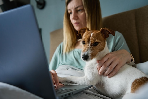 Mujer acostada en la cama con perro y usa laptop para trabajar