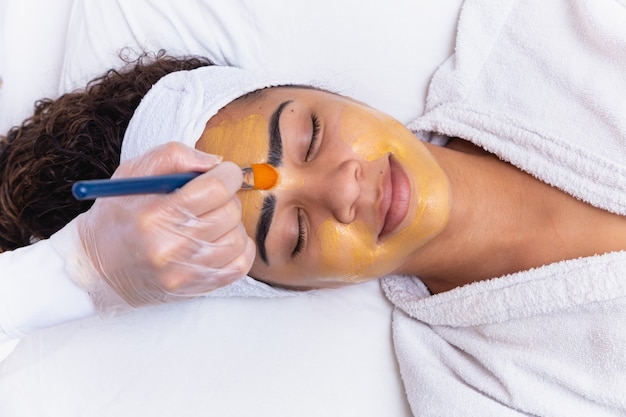 Foto la mujer está acostada en la oficina de la cosmetóloga sobre el procedimiento de hidratación de la cara. la cosmetóloga aplica una mascarilla hidratante en el rostro del paciente.