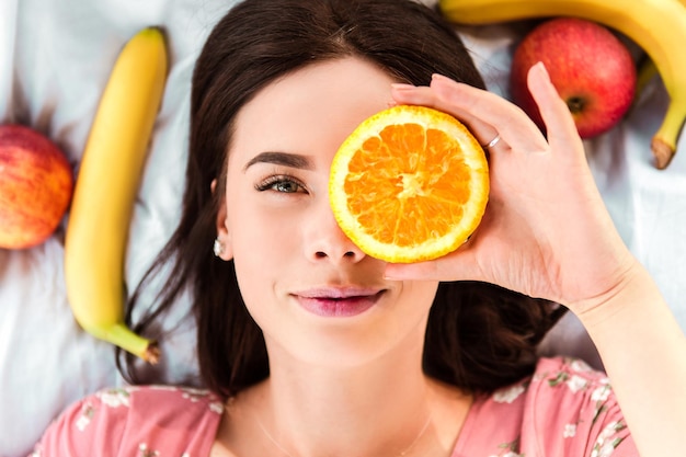 Mujer acostada sobre una sábana blanca, sonriendo y sosteniendo una rodaja de naranja