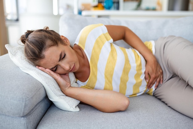 Mujer acostada en el sofá con aspecto enfermo en la sala de estar Hermosa mujer joven acostada en la cama y tomándose las manos sobre el estómago Mujer con dolor de estómago doloroso en la cama Período menstrual