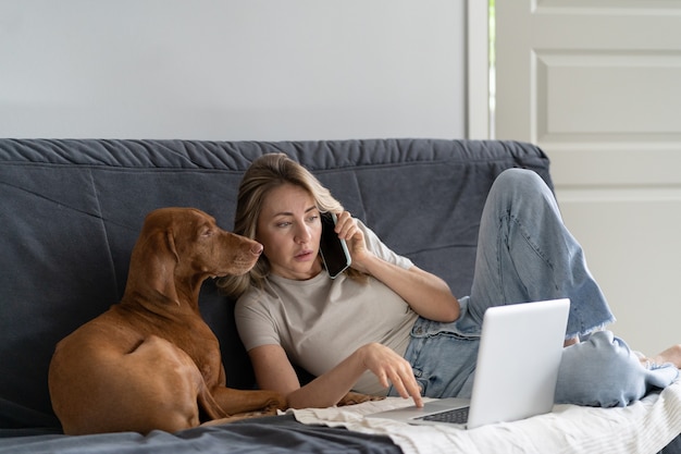Mujer acostada en el sofá con su adorable perro Vizsla, hablando por teléfono celular, trabajando de forma remota en la computadora
