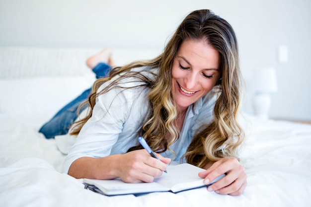 Mujer acostada en su cama, sonriendo y escribiendo en un libro con un bolígrafo