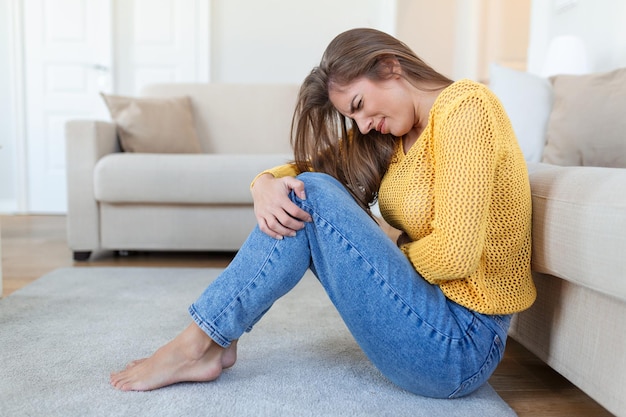 Mujer acostada en el suelo con aspecto enfermo en la sala de estar Hermosa mujer joven acostada en la cama y tomándose las manos sobre el estómago Mujer con dolor de estómago doloroso en la cama Período menstrual