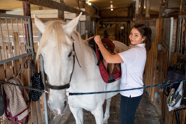 Mujer activa joven que pone la silla en la parte posterior del caballo de carreras de pan puro blanco mientras se prepara para la carrera o el entrenamiento