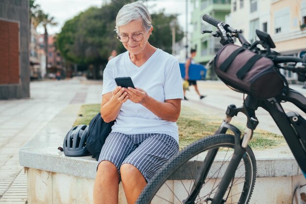 Mujer activa mayor sentada al aire libre en la ciudad cerca de su bicicleta eléctrica usando teléfono móvil