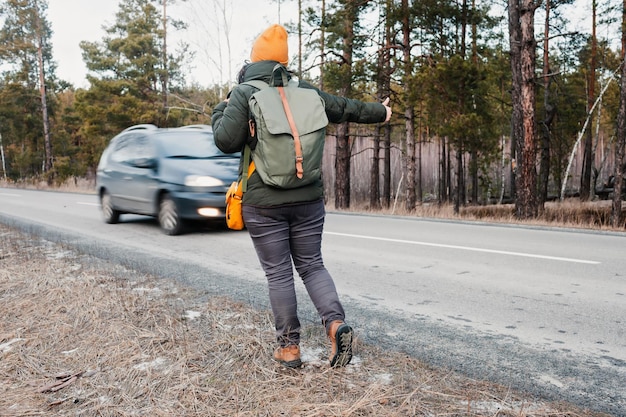 Mujer activa de mediana edad con mochila caminando en la carretera y gesto tratando de detener un camión, fondo forestal, concepto de viaje de invierno, estilo de vida saludable, turismo en solitario haciendo autostop