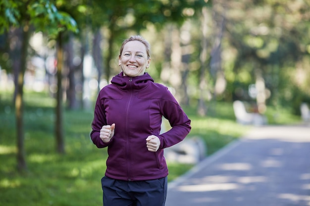 Una mujer activa de mediana edad con ropa deportiva disfruta de una carrera en un exuberante parque verde sonriendo mientras corre
