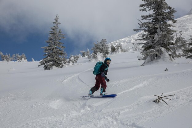 Una mujer activa practicando freeriding en una tabla de snowboard en un terreno alpino de travesía entre abetos nevados en montañas blancas