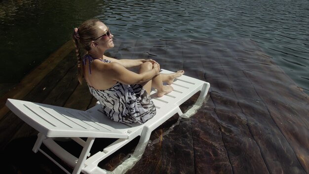 La mujer se acuesta en una tumbona con gafas de sol y un chal de seda boho La niña descansa en un muelle submarino de madera inundada El pavimento está cubierto de agua en el lago En el fondo hay una montaña y un bosque