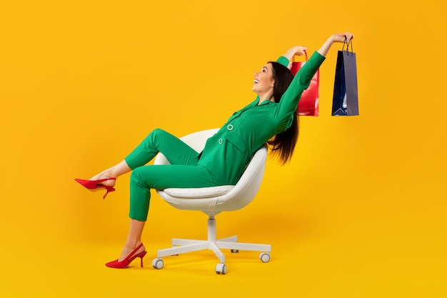 Mujer adicta a las compras feliz con muchas bolsas de compras coloridas sentada en una silla sobre fondo de estudio amarillo