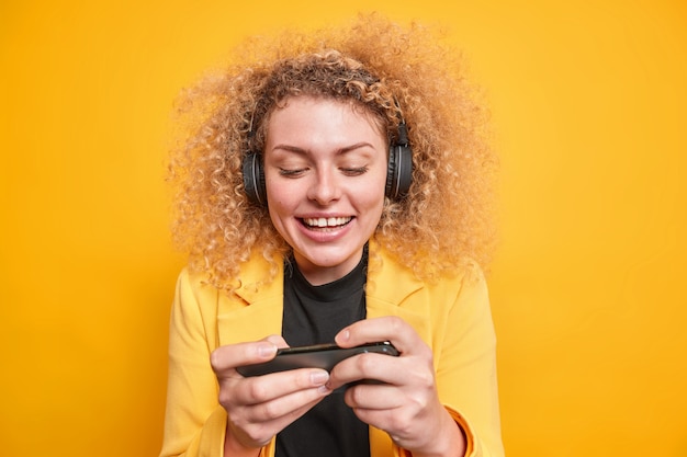 Mujer adicta a las tecnologías modernas juega videojuegos tiene smartphone horizontalmente lleva auriculares inalámbricos vestidos con chaqueta