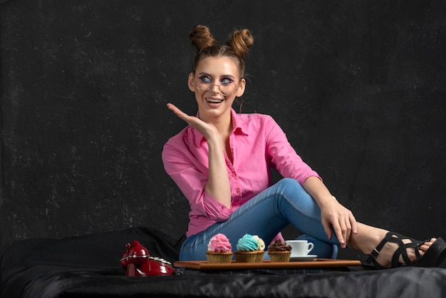 Mujer admira cupcakes con crema multicolor Hermosa chica con gafas en camisa rosa se sienta sobre fondo negro