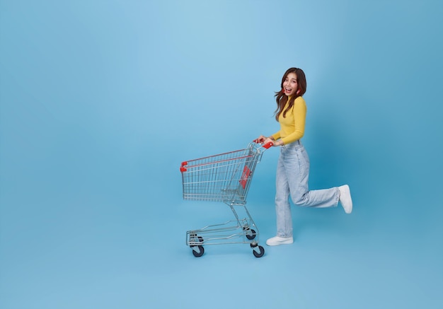 Mujer adolescente asiática feliz de larga duración empujando un carrito de compras vacío o un tranvía de compras corriendo promoción supermercado aislado en fondo azul