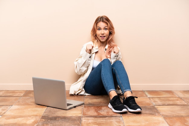 Mujer adolescente con una computadora portátil sentada en el suelo en el interior celebrando una victoria en la posición ganadora