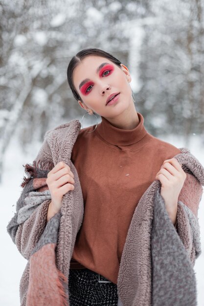 Mujer adolescente glamorosa de moda con maquillaje de color en ropa elegante vintage con suéter beige y abrigo posando en un parque con nieve. Estilo creativo de invierno femenino