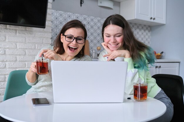 Foto mujer y adolescente sentados en casa en la cocina, bebiendo té juntos y mirando el monitor de la computadora portátil