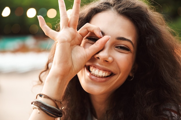 Foto mujer adolescente vivaz sonriendo y mirándote a través del signo de ok, durante la caminata en el parque verde