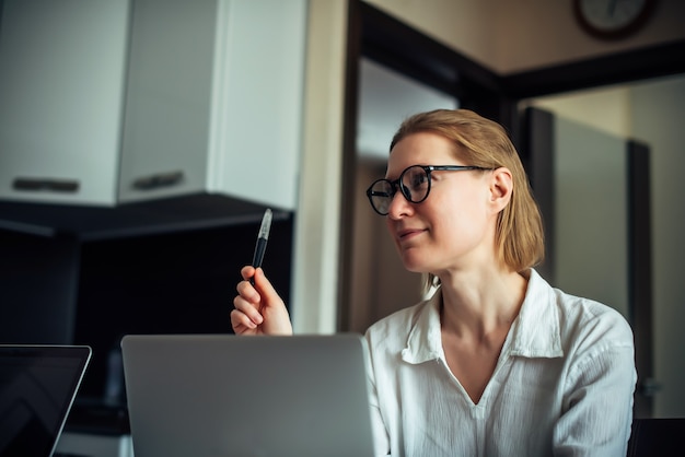 Mujer adulta de 40 años con gafas y camisa blanca está trabajando con un portátil en una oficina en casa. Escritora positiva. Freelance, coworking, trabajo a distancia, autoempleo, encierro.