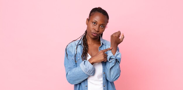 Mujer adulta afro negra que parece impaciente y enojada, apuntando al reloj, pidiendo puntualidad, quiere llegar a tiempo