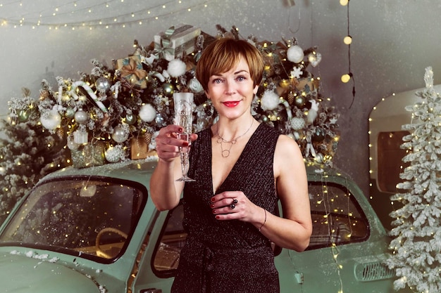 Mujer adulta bebe champán en un coche antiguo junto a un árbol de Navidad en la víspera de Año Nuevo