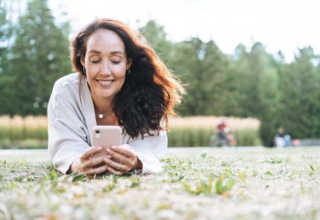 Foto mujer adulta de cabello rizado sonriente en casual usando teléfono móvil en el parque de la ciudad