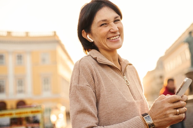 Mujer adulta caucásica sonriente que recibe un mensaje agradable de familiares por teléfono mientras está de pie al aire libre Concepto de emociones positivas