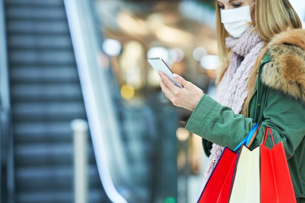 mujer adulta de compras en el centro comercial con un teléfono inteligente con una máscara, concepto de coronavirus