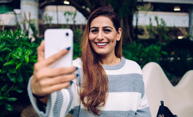 Mujer adulta contenta con cabello largo en suéter casual a rayas tomando fotos con teléfono inteligente