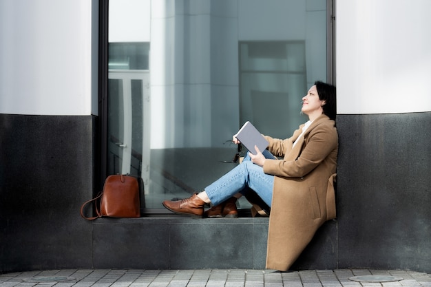 Una mujer adulta elegantemente vestida se sienta en la calle en el alféizar de la ventana de un edificio leyendo un libro