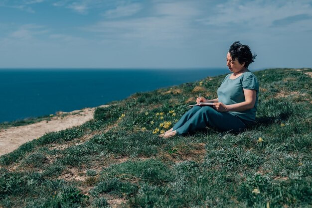 Mujer adulta escribe cuidadosamente el diario de planes en un día soleado en una montaña contra el fondo del mar