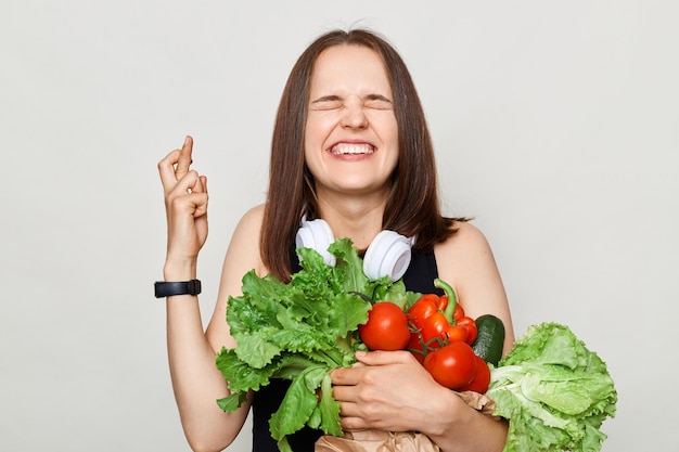 Mujer adulta esperanzada con cabello castaño aislada sobre fondo blanco sosteniendo vegetales cruzando su dedo cerrando los ojos pidiendo deseo orando por buena suerte