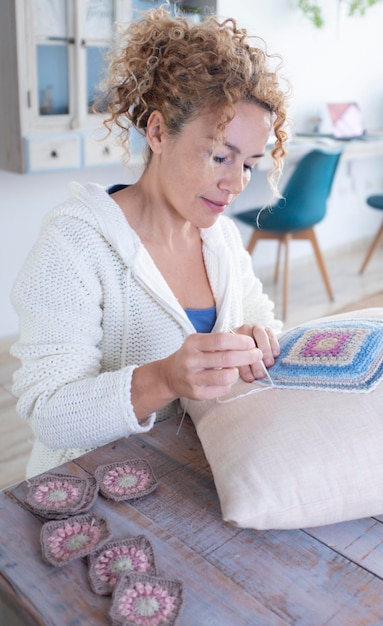 Mujer adulta feliz cosiendo una almohada con cuadrados de ganchillo hechos a mano sentada cómodamente en la mesa en casa Gente interior ocio hobby actividad concepto estilo de vida Señora sonriente trabajando sola en casa