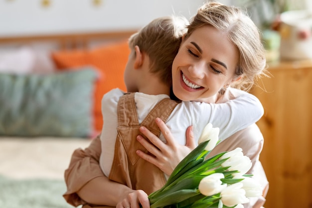 Mujer adulta feliz con tulipanes sonriendo con los ojos cerrados y abrazando a un niño en agradecimiento mientras celebra las vacaciones en casa