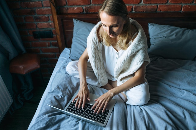 Mujer adulta independiente de cuarenta años con cabello largo rubio en ropa casual usando una computadora portátil sentada en la cama en casa vista desde arriba