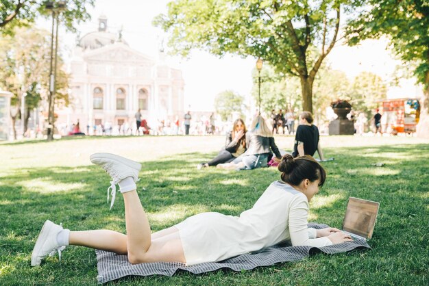 Mujer adulta joven acostada con una computadora portátil en el parque de la ciudad sobre hierba verde