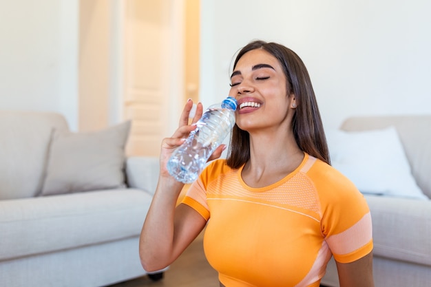 Mujer adulta joven bebiendo agua, sentada en una colchoneta de fitness y descansando después de entrenar en casa