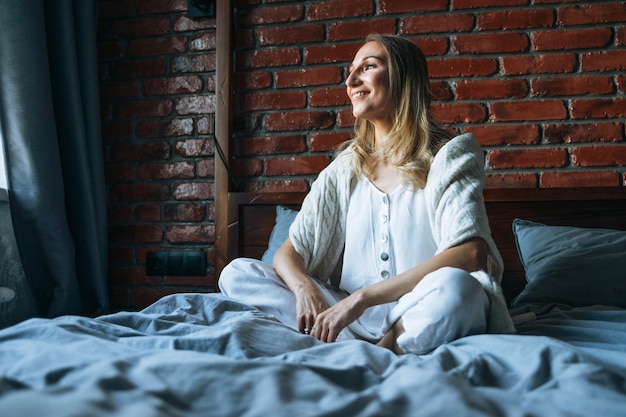 Mujer adulta joven con cabello largo rubio en ropa casual sentada en la cama en casa