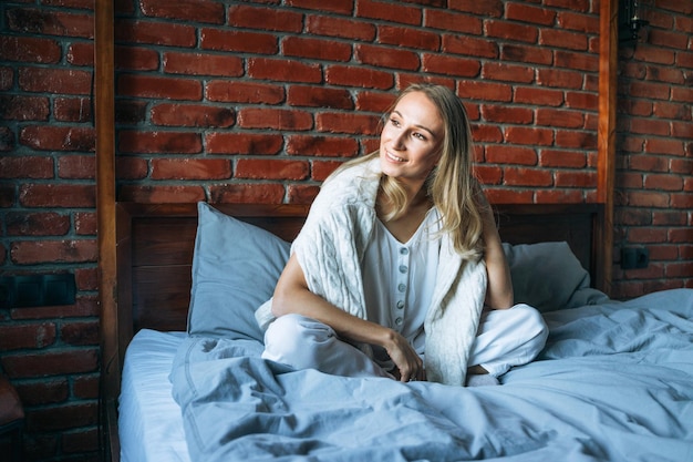 Mujer adulta joven con cabello largo rubio en ropa casual sentada en la cama en el hogar