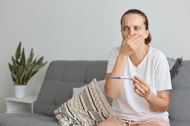 Mujer adulta joven embarazada con camiseta blanca con prueba de embarazo en las manos, sentada en el sofá en casa, cubriéndose la boca con las manos, se siente mal, tiene toxicosis y náuseas.